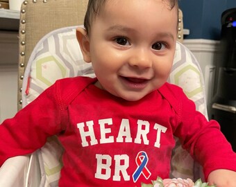 May include: A baby wearing a red long-sleeved shirt with the text "HEART BR" and a blue and red ribbon. The baby is sitting in a high chair.