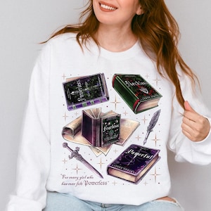 Może przedstawiać: Biała bluza z nadrukiem książek, miecza i pióra. Książki są w odcieniach fioletu i zieleni, z widocznymi słowami "Fearless" i "Powerful". Widoczny jest również tekst "For every girl who has ever felt powerless".