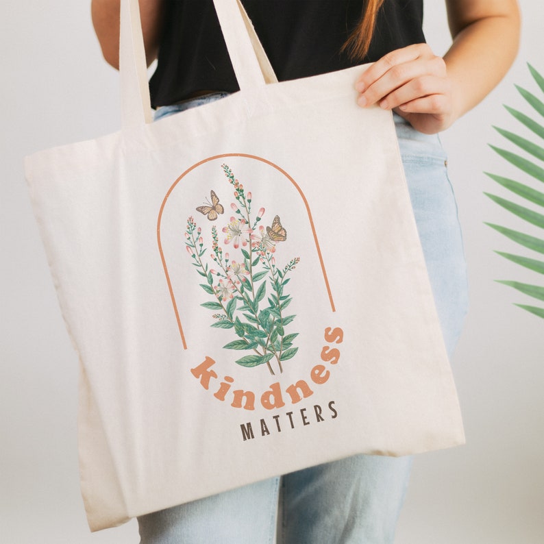 May include: Cream-colored tote bag with a floral and butterfly design inside a brown arch. The words "kindness matters" are printed in brown below the design. The bag has two cream-colored handles.