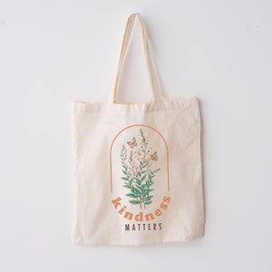May include: Cream-colored tote bag with a floral design inside a brown arch. The design includes butterflies, flowers, and the words "kindness matters" in an orange font. The bag has long handles.
