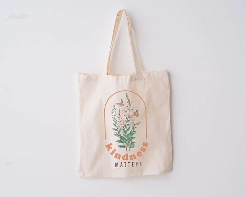 May include: Cream-colored tote bag with a floral design inside a brown arch. The design includes butterflies, flowers, and the words "kindness matters" in an orange font. The bag has long handles.