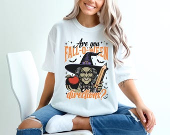 May include: White t-shirt featuring a Halloween-themed graphic. The design includes a witch holding an apple and a pencil, with the text "Are you Fall-o-ween directions?" in orange and black.