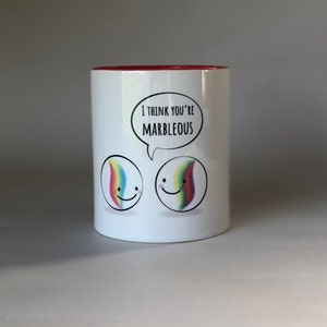 May include: White ceramic coffee mug with a red rim. The mug features a cartoon image of two marbles with smiling faces and rainbow swirls. The text "I think you're MARBLEOUS" is written in a speech bubble above the marbles.