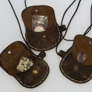 May include: Three small, open, brown leather pouches with black cords. Each pouch contains a different item: a wrapped candy, a speckled stone, and a purple crystal. The pouches are stitched with white thread and have metal closures.