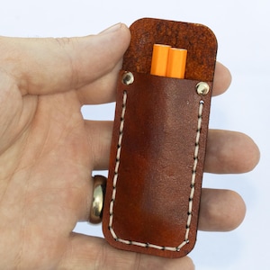 May include: A brown leather pencil case with white stitching and two orange pencils inside. The case is held in a hand against a white background. The case has a rectangular shape with rounded edges and is secured with two rivets.