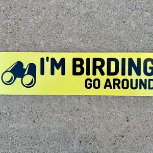 Op de afbeelding: Gele sticker met zwarte tekst en een zwart silhouet van een verrekijker. De tekst luidt "I'm Birding Go Around".