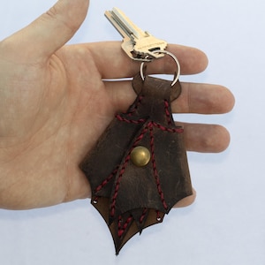May include: A brown leather keychain in a bat shape, with red stitching and a brass button. The keychain is attached to a silver key ring with keys.