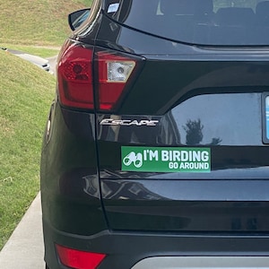 Op de afbeelding: Een zwarte Ford Escape met een groene bumper sticker waarop staat "I'm Birding Go Around" met een afbeelding van een verrekijker.