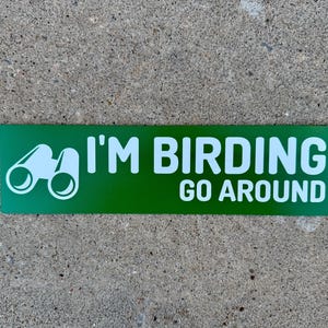 Op de afbeelding: Groene rechthoekige sticker met witte tekst die "I'm Birding Go Around" zegt en een wit silhouet van een verrekijker.