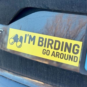 Op de afbeelding: Een gele bumper sticker met zwarte tekst die "I'm Birding Go Around" luidt. Links van de tekst is een verrekijker afgebeeld.