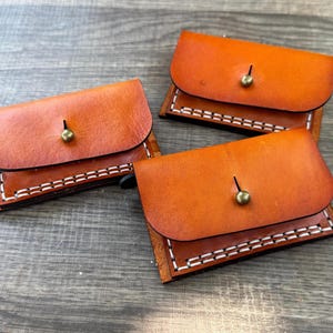 May include: Three small, handcrafted leather wallets in shades of brown and tan. Each wallet features a rounded flap secured with a brass-colored stud. The edges are hand-stitched with white thread, creating a contrasting border. The wallets are arranged on a weathered wooden surface.
