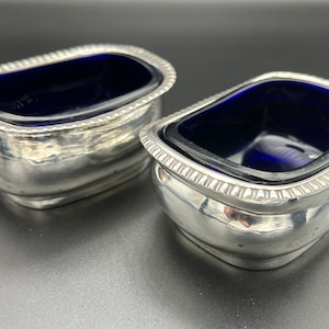 May include: Two silver salt cellars with blue glass inserts. The salt cellars are oval shaped and have a beaded rim.