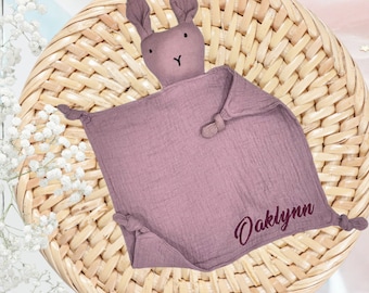 Può includere: Una morbida copertina per neonati in mussola di colore malva con una testa di coniglio di peluche attaccata. La copertina ha un angolo annodato e il nome "Oaklynn" ricamato in filo marrone.