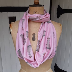 May include: Pink infinity scarf with a repeating pattern of grey and white illustrations of women wearing dresses.