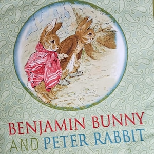 May include: A square fabric featuring an illustration of two rabbits, one wearing a red scarf. The image is set within a blue circle on a light green background with a paisley pattern. Text at the bottom reads "BENJAMIN BUNNY AND PETER RABBIT."