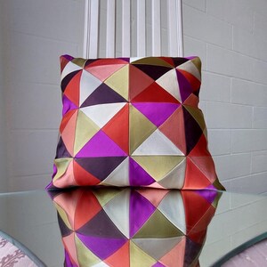 May include: A geometric patterned pillow with a mix of red, pink, purple, green, and black triangles. The pillow is sitting on a glass table.