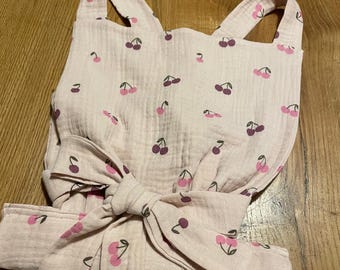 May include: A light pink baby bib with a cherry pattern. The bib has a bow in the front and straps that go over the shoulders. The cherries are in shades of pink and purple.