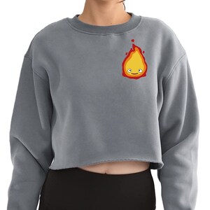 a woman wearing a grey long sleeve crop top with a flame with layers of red, orange, and yellow. There are white eyes and a red smile making up the face.