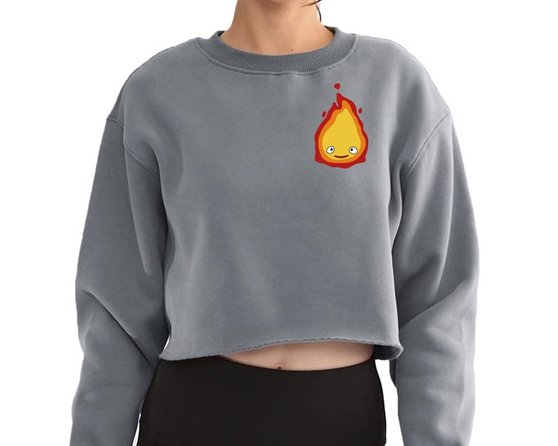a woman wearing a grey long sleeve crop top with a flame with layers of red, orange, and yellow. There are white eyes and a red smile making up the face.