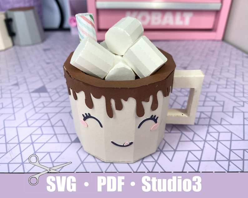 a tan mug with a smiling face on it. There is chocolate dripping over the top edge. There are also paper marshmallows sitting inside the mug with a stir stick. The bottom shows that it is a SVG, PDG, and Studio3 file.