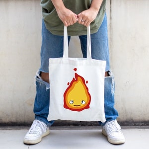 a white tote bad with a flame with layers of red, orange, and yellow. There are white eyes and a red smile making up the face.