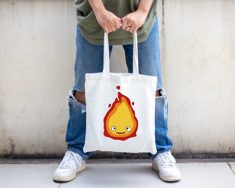 a white tote bad with a flame with layers of red, orange, and yellow. There are white eyes and a red smile making up the face.