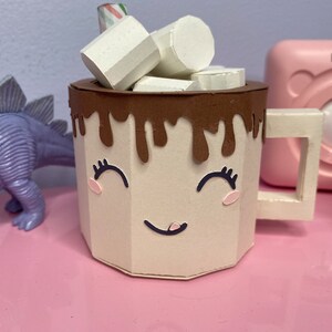 a tan mug with a smiling face on it. There is chocolate dripping over the top edge. There are also paper marshmallows sitting inside the mug with a stir stick.