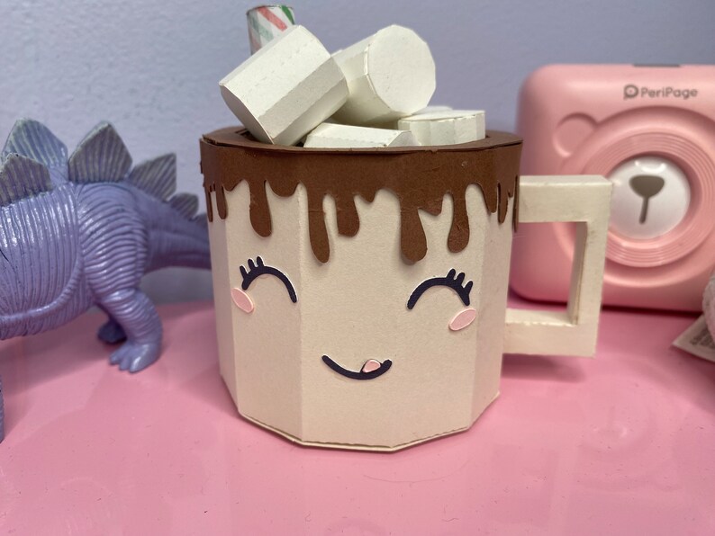 a tan mug with a smiling face on it. There is chocolate dripping over the top edge. There are also paper marshmallows sitting inside the mug with a stir stick.