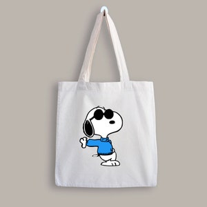 K&ouml;nnte beinhalten: Wei&szlig;e Stofftasche mit einer Schwarzwei&szlig;-Illustration von Snoopy, der eine Sonnenbrille und einen blauen Pullover tr&auml;gt.