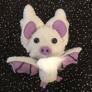 以下が含まれることがあります： 紫色の耳と羽根を持つ灰色のフェルトの蝙蝠。蝙蝠は黒い目と紫色の鼻をしています。蝙蝠は白いふわふわの尻尾を持っています。