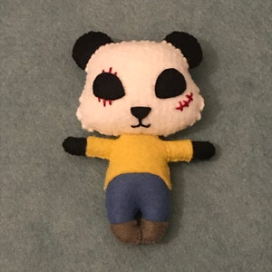 Puede incluir: Un juguete de peluche de panda hecho a mano con pelaje blanco y negro, que lleva una camisa amarilla y pantalones vaqueros azules. El panda tiene ojos negros cosidos y una boca roja cosida.
