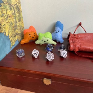 May include: A wooden box with a red stain and a brass latch. The box contains six dice and four felt toys shaped like monsters. The toys are orange, green, and blue. The dice are black and white with numbers on each side.