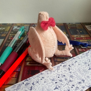 May include: A pink felt bird with a red bow tie. The bird is sitting on a bookshelf with a blue bookmark.