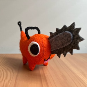May include: An orange felt plush toy shaped like a creature with a chainsaw head. The toy has a black handle, black eyes, and a small pink mouth. The chainsaw is gray.