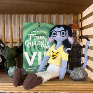 Può includere: Una bambola di feltro con la pelle blu, che indossa una maglietta gialla con un'ape, occhiali e una treccia scura. È seduta davanti a una carta verde con la scritta "Dimension 20 Time Quangle Live". Sono visibili anche una piccola bottiglia di vetro e una pietra.