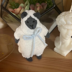 Pode incluir: Um pequeno brinquedo de pelúcia de carneiro branco com orelhas pretas e uma fita azul amarrada ao redor do pescoço.
