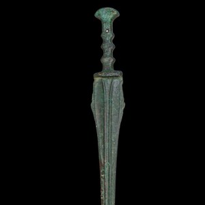 May include: A bronze dagger with a long, slender blade and a detailed handle. The handle features a bulbous top and a series of decorative ridges. The blade has a central ridge and is flanked by parallel grooves. The dagger has a weathered, antique appearance.