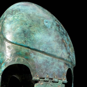 May include: A bronze helmet with a green patina. The helmet is made of two pieces, the top and the bottom, which are joined together by a hinge. The helmet has a rounded top and a flat bottom. The helmet is missing a piece on the right side.