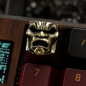 May include: A gold-coloured metal keycap featuring a detailed, angry-looking face mask. The keycap is on a red and black keyboard.