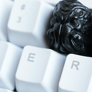 Puede incluir: Una tecla negra esculpida con un diseño detallado de rostro, ubicada entre teclas blancas de teclado. Esta tecla personalizada reemplaza una tecla estándar, agregando una estética única a una configuración informática.
