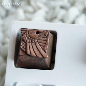 May include: A copper-colored keycap with a detailed design of a winged creature. The keycap is in a white plastic keycap holder.