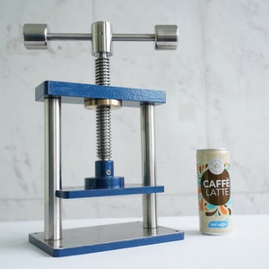 Könnte beinhalten: Eine blaue und silberne Metallpresse mit einem Schraubmechanismus und einem Griff. Die Presse steht auf einer weißen Oberfläche neben einer Dose Caffè Latte.