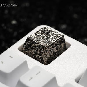 May include: A silver metal keycap with a textured surface resembling the moon. The keycap is sitting on a white keyboard.