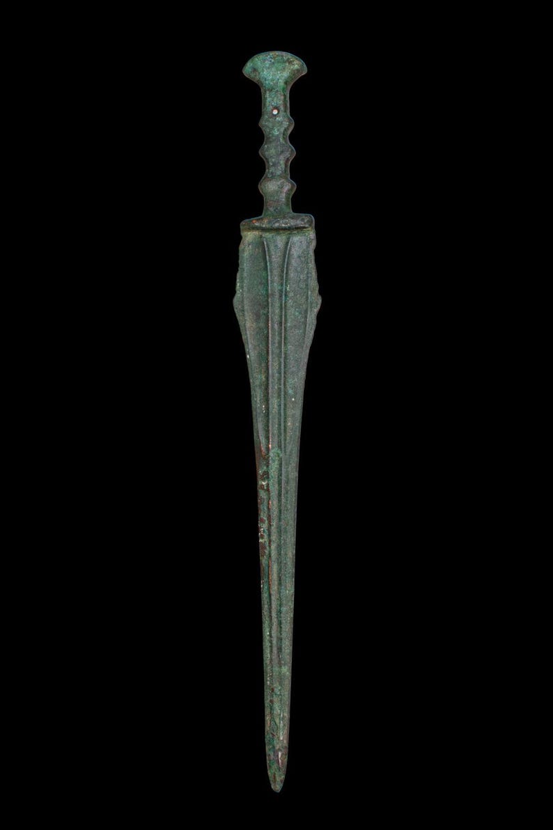 May include: A bronze dagger with a long, slender blade and a detailed handle. The handle features a bulbous top and a series of decorative ridges. The blade has a central ridge and is flanked by parallel grooves. The dagger has a weathered, antique appearance.