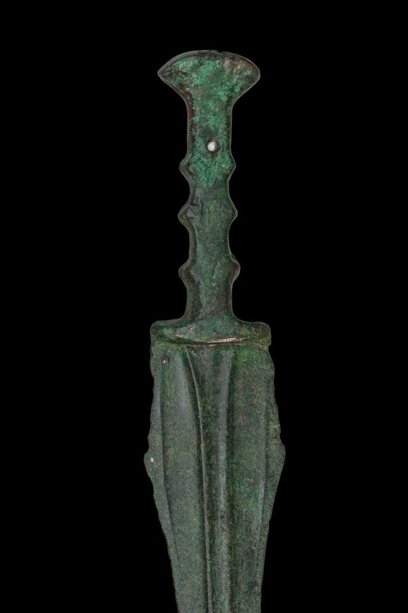 May include: A detailed image of an antique bronze dagger with a green patina. The dagger features a flared handle, a ridged grip, and a leaf-shaped blade. The background is solid black.