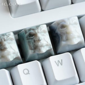 Può includere: Primo piano di tasti per tastiera personalizzati con volti scolpiti. I tasti sono un mix di blu, bianco e grigio, con un aspetto dettagliato e invecchiato. I tasti sono disposti su una tastiera bianca, con il sito web KEYRELIC.COM visibile.