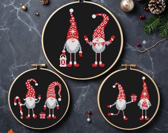May include: Three black embroidery hoops with cross-stitched Christmas gnomes. The gnomes have red and white hats, gray beards, and hold gifts and a lantern. The text "CrossStitchFoxy" is at the bottom.