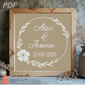 Può includere: Un'opera d'arte a punto croce incorniciata con i nomi "Alex & Jessica" e la data "12-09-2026" in scrittura bianca. I nomi sono racchiusi da una ghirlanda floreale bianca su sfondo beige, all'interno di una cornice di legno.