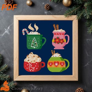 Può includere: Schema a punto croce con quattro tazze di bevande calde con panna montata e decorazioni festive. Le tazze sono verdi, rosa, rosse e verdi, ognuna con disegni unici come un albero di Natale, stelle di Natale, una renna e cuori. L'opera è incorniciata.