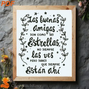 May include: A framed cross-stitch artwork with the text "Las buenas Amigas son como las Estrellas, no siempre las ves pero sabes que siempre están ahí" in black on a white background. The frame is made of light brown wood.